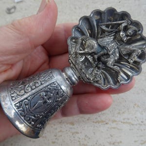 May include: A silver-coloured bell-shaped object with intricate embossed patterns and a decorative top featuring a detailed relief of a figure on horseback. The bell has a cross design.