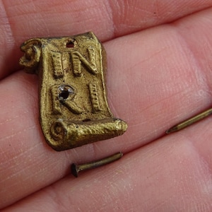 May include: A small, aged brass scroll pendant with the letters INRI and two small brass nails. The pendant has a hole at the top for hanging. The items are displayed on a skin-toned background.