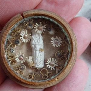 Puede incluir: Pequeño colgante religioso redondo y antiguo con un interior detallado. El colgante presenta una estatua blanca rodeada de pequeños detalles florales blancos y dorados. El marco es de madera y tiene un color marrón claro.