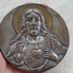 May include: A silver and gold toned metal medallion depicting Jesus Christ with a heart on his chest. The heart is surrounded by a crown of thorns and has three nails in it. The medallion is oval shaped and has a raised rim.