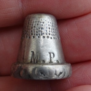May include: A silver thimble with the initials "M.P." engraved on the side. The thimble has a textured top and a decorative floral pattern around the base. This sewing tool is designed to protect the finger while sewing.