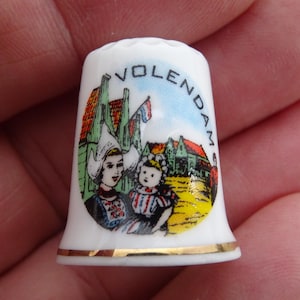 May include: White porcelain thimble with a gold trim. The thimble features a colorful illustration of a Dutch village scene with two girls in traditional clothing. The text "VOLENDAM" is printed in a blue circle around the top of the thimble.