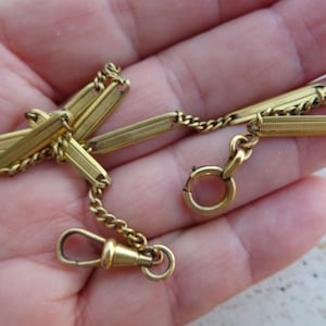 Può includere: Una catena per orologio in metallo color oro caratterizzata da una serie di maglie rettangolari testurizzate collegate da piccole catene a maglia barbazzale. L'oggetto include una chiusura a molla circolare a un'estremità e un moschettone girevole.