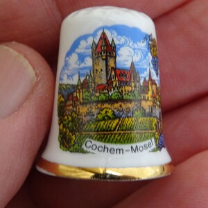 May include: White porcelain thimble with a colorful illustration of a castle and vineyards. The thimble has a gold trim and the text "Cochem-Mosel" printed on the bottom.
