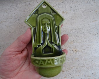 May include: A green ceramic wall plaque with a raised image of a woman in prayer. The plaque has a small, recessed dish at the bottom with the word "AME" inscribed.