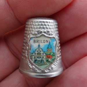 Op de afbeelding: Een zilveren vingerhoed met een gedetailleerd schildontwerp met het woord "BRILON" en een kleurrijke illustratie van een stadsgezicht. De vingerhoed heeft een gestructureerd oppervlak en een decoratieve rand.