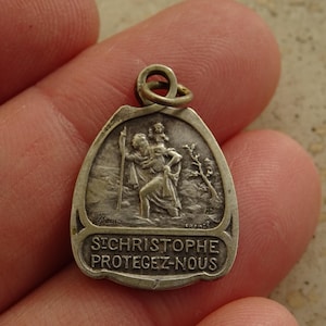 Könnte beinhalten: Ein silberfarbener Metallanhänger mit einem Relief des heiligen Christophorus, der ein Kind auf der Schulter trägt. Das Design zeigt einen Stab, einen Baum und den geprägten französischen Text "ST CHRISTOPHE PROTEGEZ-NOUS" unten. Mit runder Öse.