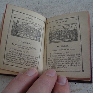 May include: An open book with black and white illustrations depicting scenes from the Stations of the Cross. The book has a red spine and is open to pages 74 and 75. The text on the pages is in French.