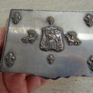 Op de afbeelding: Een rechthoekige zilveren doos met sierlijke details. De doos heeft een centraal reliëf van een figuur en twee kleinere hondachtige figuren, met fleur-de-lis accenten. De doos wordt in een hand gehouden.