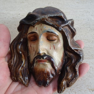 May include: A wooden sculpture of a man's face with a crown of thorns, closed eyes, and a beard. The face is painted in shades of brown, white, and red.