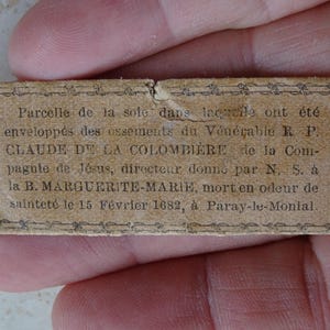 Può includere: Un piccolo cartellino rettangolare di carta marrone con testo nero. Il testo recita: "Parcelle de la sole dans laczaile ont été enveloppés des ossements du Vénérable RP. CLAUDE DE LA COLOMBIERE de la Com- pagule de Jésus, directeur donne par N. S. a la B. MARGUERITE-MARIE, morten odeur de sainteté le 15 Février 1682, à Paray-le-Monial."