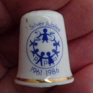 Puede incluir: Dedal de cerámica blanca con borde dorado y texto y gráficos azules. El dedal está decorado con las palabras "Silver Jubilee" y los años "1961-1986". El diseño incluye un círculo de figuras estilizadas.