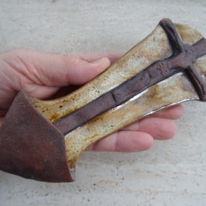 May include: A ceramic wall hanging in the shape of a cross with a brown and white glaze. The cross features a raised figure of Jesus Christ.