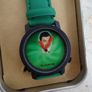 May include: Black wristwatch with a green face featuring a portrait. The watch has a black bezel and a green strap. The watch face has a green background with a portrait and red hands. The watch is in a metal tin.