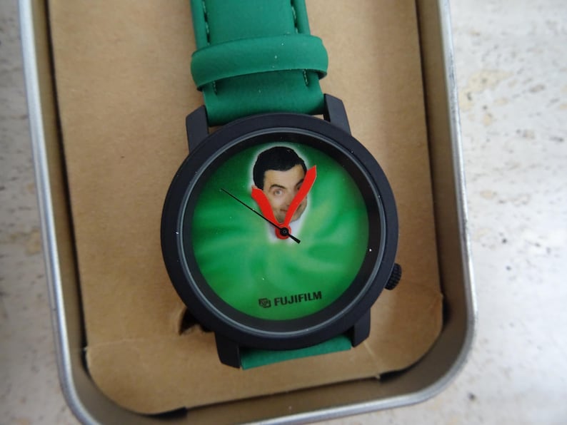 May include: Black wristwatch with a green face featuring a portrait. The watch has a black bezel and a green strap. The watch face has a green background with a portrait and red hands. The watch is in a metal tin.