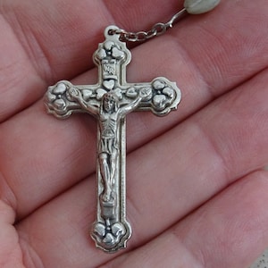 Könnte beinhalten: Ein silberner Rosenkranz mit einem detaillierten Kruzifix. Das Kruzifix zeigt eine Darstellung von Jesus Christus und ist mit dekorativen floralen Elementen verziert. Der Rosenkranz wird in einer Hand gehalten und zeigt das komplizierte Design und die religiöse Bedeutung.