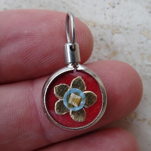 Pode incluir: Pingente redondo prateado com fundo vermelho. Apresenta um design floral dourado com um centro azul e branco. Possui uma pequena argola na parte superior para pendurar.