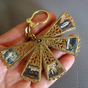 Pode incluir: Um porta-chaves de metal dourado com um design em forma de cruz. A cruz apresenta seis secções em forma de leque, cada uma contendo uma pequena imagem. O porta-chaves tem um design ornamentado e vazado e um fecho.
