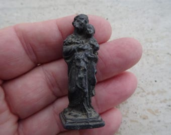 May include: A small, black metal figurine of a man holding a child. The figurine is standing on a square base.