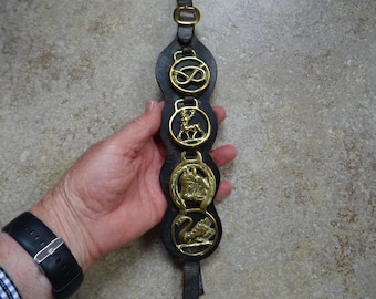 May include: A black leather horse harness decoration with four brass medallions. The medallions feature a knot, a deer, a horse and rider, and a swan. The harness has a leather strap and buckle.