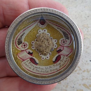 Pode incluir: Um relicário redondo prateado com uma tampa de vidro. O relicário contém um pequeno pedaço de tecido com uma relíquia marrom no centro. O tecido é decorado com tinta dourada e vermelha e tem o texto "S. Florentia V.M. Filia Duci Sociis S. Urbis. Vixy. Martyre".