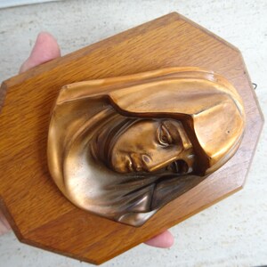May include: A bronze-toned relief sculpture of a woman's face, mounted on a wooden, octagonal plaque. The sculpture depicts a serene face with closed eyes and draped head covering. The wood has a warm, natural finish.