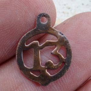 May include: A small, circular, bronze-colored pendant with the number 13 cut out in the center. The pendant has a small loop at the top for attaching to a chain or cord. The edges of the pendant are slightly worn.