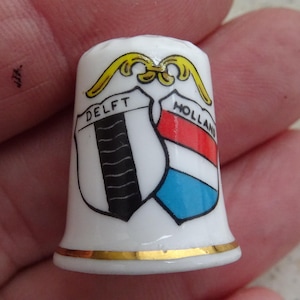 Può includere: Un ditale in ceramica bianca con finiture dorate. Il ditale presenta due scudi, uno con la scritta "DELFT" e un disegno in bianco e nero, e l'altro con "HOLLAND" e un disegno rosso, bianco e blu. Un elemento decorativo dorato è in cima.