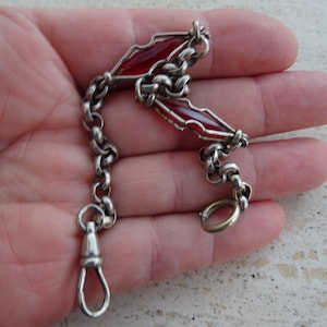 May include: A silver-toned metal chain featuring two elongated, diamond-shaped links with translucent red inserts. The chain includes a circular jump ring at one end and a swivel lobster-style clasp at the other, connected by a series of round links.