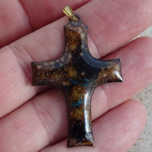 Pode incluir: Um pingente de cruz de cerâmica marrom e preto com detalhes dourados. A cruz tem um padrão de mármore com manchas azuis e douradas.