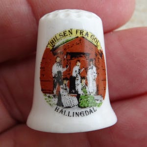 May include: White ceramic thimble with a detailed illustration of a building and figures. The thimble has the text "HILSEN FRA GOL" and "HALLINGDAL" printed on it. A collectible item.