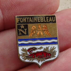 Könnte beinhalten: Rechteckige Vintage-Anstecknadel mit dem Wort "FONTAINEBLEAU" oben. Die Nadel hat einen goldenen Rand und enthält ein blau-weißes Design, ein rotes Tier und den Buchstaben "N".