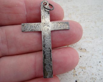 Könnte beinhalten: Silberner Kreuzanhänger mit eingraviertem Text und Daten. Das Kreuz trägt das Datum "1846" und die Inschrift "12. Juli". Der Anhänger hat oben eine kleine Schlaufe zum Aufhängen.