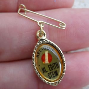 Puede incluir: Un alfiler dorado con un colgante ovalado. El colgante presenta el texto "RELIQUE" y "HL. EDITH STEIN" sobre un fondo verde con un círculo rojo y una línea roja vertical. El alfiler se sostiene en una mano.