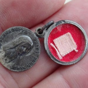 Pode incluir: Um pequeno pingente religioso redondo prateado. Um lado apresenta um relevo do rosto de uma mulher com as palavras "BERNADETTE" e "PRIEZ POUR NOUS". O outro lado abre para revelar um interior vermelho com um pequeno objeto quadrado de tecido.