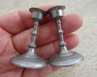 May include: Two small, antique silver candle holders. Each has a round base, a decorative stem, and a small cup to hold a candle. The candle holders are made of metal and have a weathered appearance.