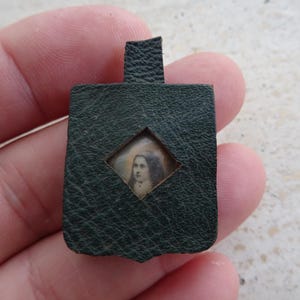 May include: A dark green textured leather pendant featuring a small diamond-shaped cutout in the center. Inside the cutout is a miniature portrait of a person with long dark hair and a halo, set against a light background, with a small loop at the top.