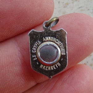 Peut inclure: Pendentif argenté en forme de bouclier, avec l'inscription "EX CRYPTA ANNUNCIATIONIS NAZARETH" autour d'un motif circulaire central. Le pendentif est doté d'une petite boucle en haut pour l'attache.