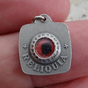 Könnte beinhalten: Silberfarbener religiöser Anhänger mit der Aufschrift "RELIQUIA". Der Anhänger hat eine quadratische Form mit einem kreisförmigen Zentrum, das ein rot-schwarzes Design enthält. Kleine Kreuzsymbole befinden sich auf beiden Seiten des Wortes.