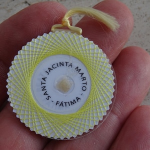 May include: A round religious pendant with a yellow thread design around a white center. The center has text that reads "SANTA JACINTA MARTO - FÁTIMA". A yellow string is attached at the top.