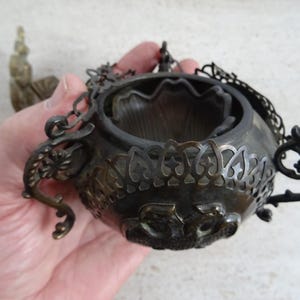 Puede incluir: Un quemador de incienso antiguo de bronce con intrincados diseños florales y geométricos. El quemador tiene forma de cuenco redondo con una cadena y un asa decorativas. Una pequeña estatua es visible en el fondo.