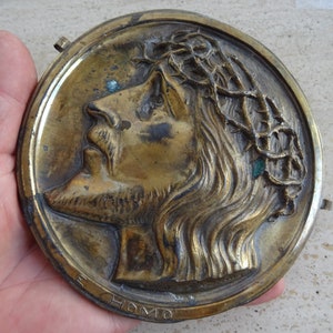 May include: A round, brass plaque with a relief sculpture of a man's face. The man is wearing a crown of thorns and has a beard. The plaque is inscribed with the words "Ecce Homo".