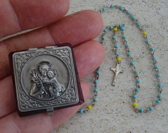 May include: A silver locket with a religious image of a man holding a child. The locket is open and has a red interior. A blue and yellow beaded rosary chain is also pictured.