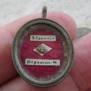 May include: A round metal pendant featuring a glass front covering a red fabric background. Inside, a small diamond-shaped paper holds a textured relic, flanked by two white paper strips with the black printed text S. Joannis and Nepomuc. M.