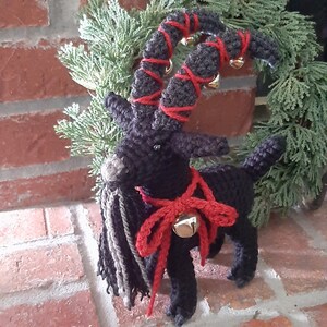 May include: A black crocheted goat with red and black yarn horns and a red ribbon with a silver bell. The goat is standing on a brick surface.