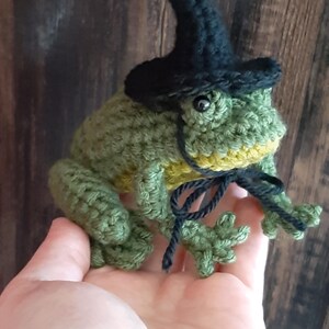 May include: A handmade crocheted frog, predominantly green with yellow accents, wearing a small black witch's hat. The frog is holding a black string. The craftwork is detailed and whimsical.