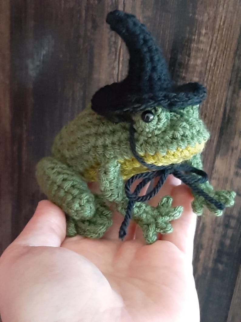 May include: A handmade crocheted frog, predominantly green with yellow accents, wearing a small black witch's hat. The frog is holding a black string. The craftwork is detailed and whimsical.