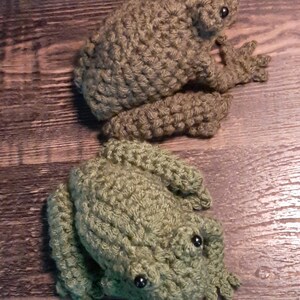 May include: Two handmade crochet frogs in shades of green and brown. The frogs have detailed features, including bulging eyes and textured bodies. The frogs are positioned on a wooden surface, showcasing the craftsmanship of the crochet work.