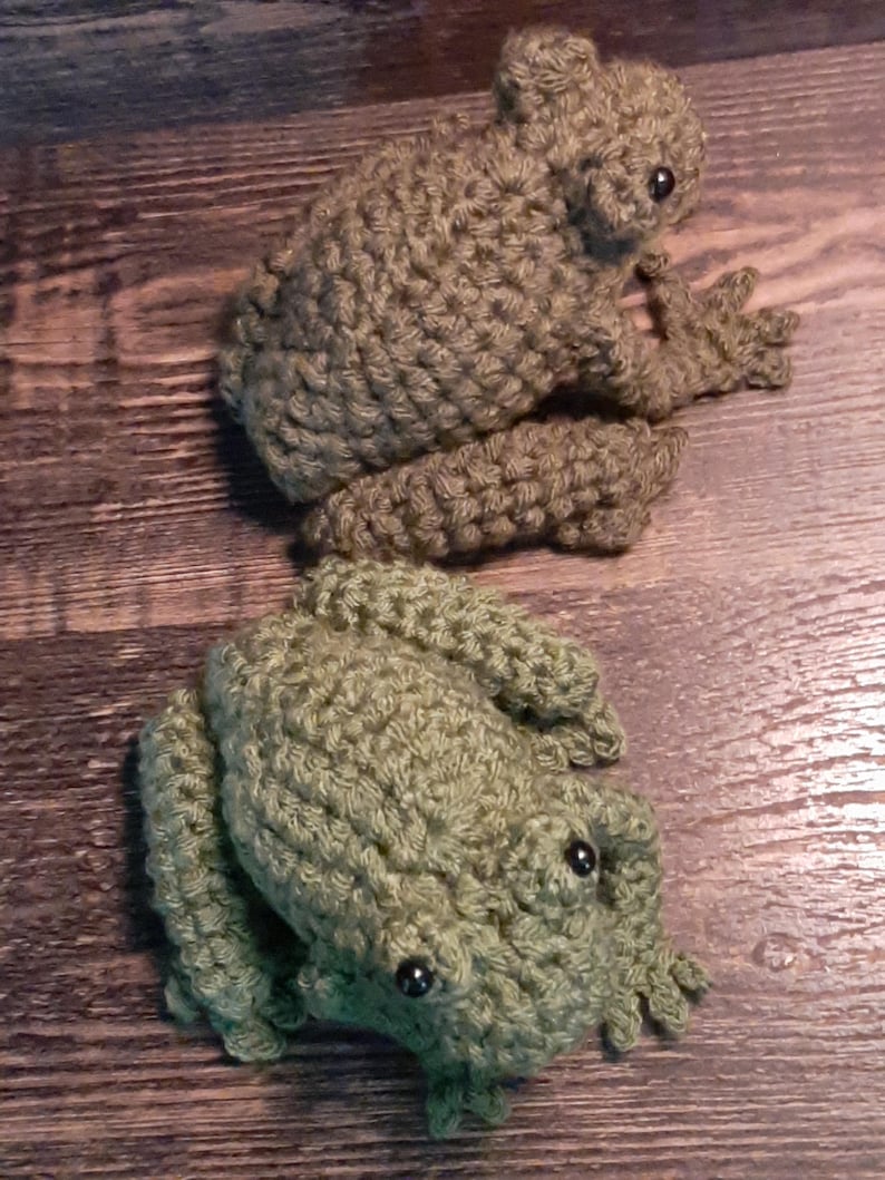 May include: Two handmade crochet frogs in shades of green and brown. The frogs have detailed features, including bulging eyes and textured bodies. The frogs are positioned on a wooden surface, showcasing the craftsmanship of the crochet work.