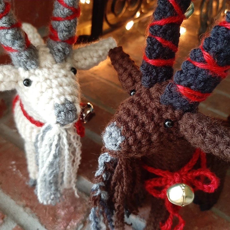 May include: Two crocheted goat ornaments with red and white striped horns. The goats are wearing red and white scarves and have bells attached to their necks. One goat is white with a gray beard and the other is brown with a gray beard.